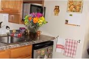 A small kitchen area with wooden cabinets, a countertop with a vase of colorful flowers, a microwave, and a refrigerator with magnets. On the wall, there is a calendar, a clock, and two hanging towels.