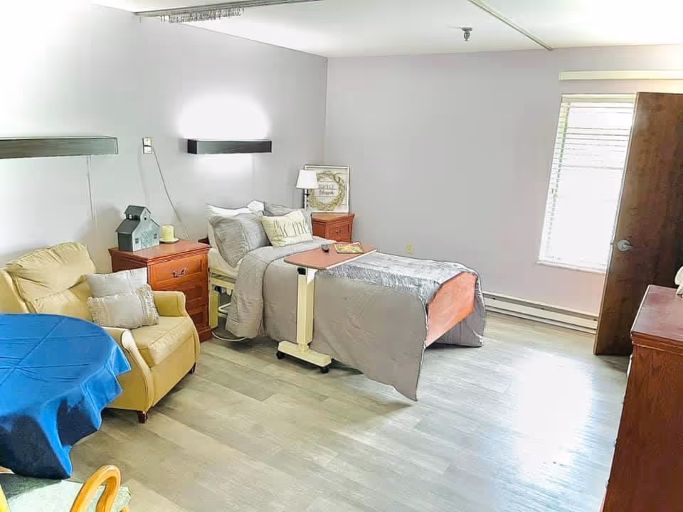 A bright, clean bedroom in a skilled nursing facility with a single bed covered in gray bedding, a small overbed table, two wooden nightstands with lamps and decor, a comfortable beige armchair with a pillow, a round table covered with a blue tablecloth, and a window with blinds letting in natural light.