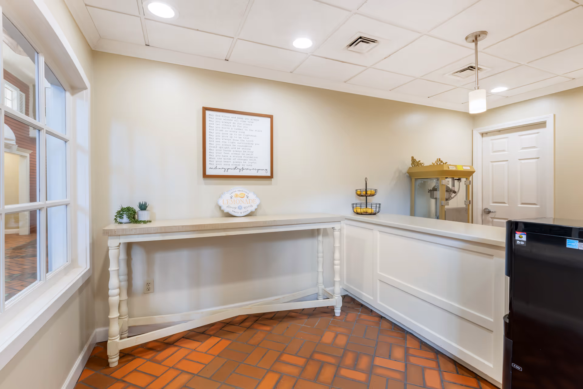 Interior corner of a room with a white wooden table against a beige wall, decorated with small potted plants and a decorative lemonade sign. Above the table is a framed text artwork. To the right is a white counter with a two-tiered fruit basket holding lemons and a vintage-style popcorn machine. The floor is covered with reddish-brown tiles, and the ceiling has recessed lighting and a hanging pendant light. A black refrigerator is partially visible on the right side.
