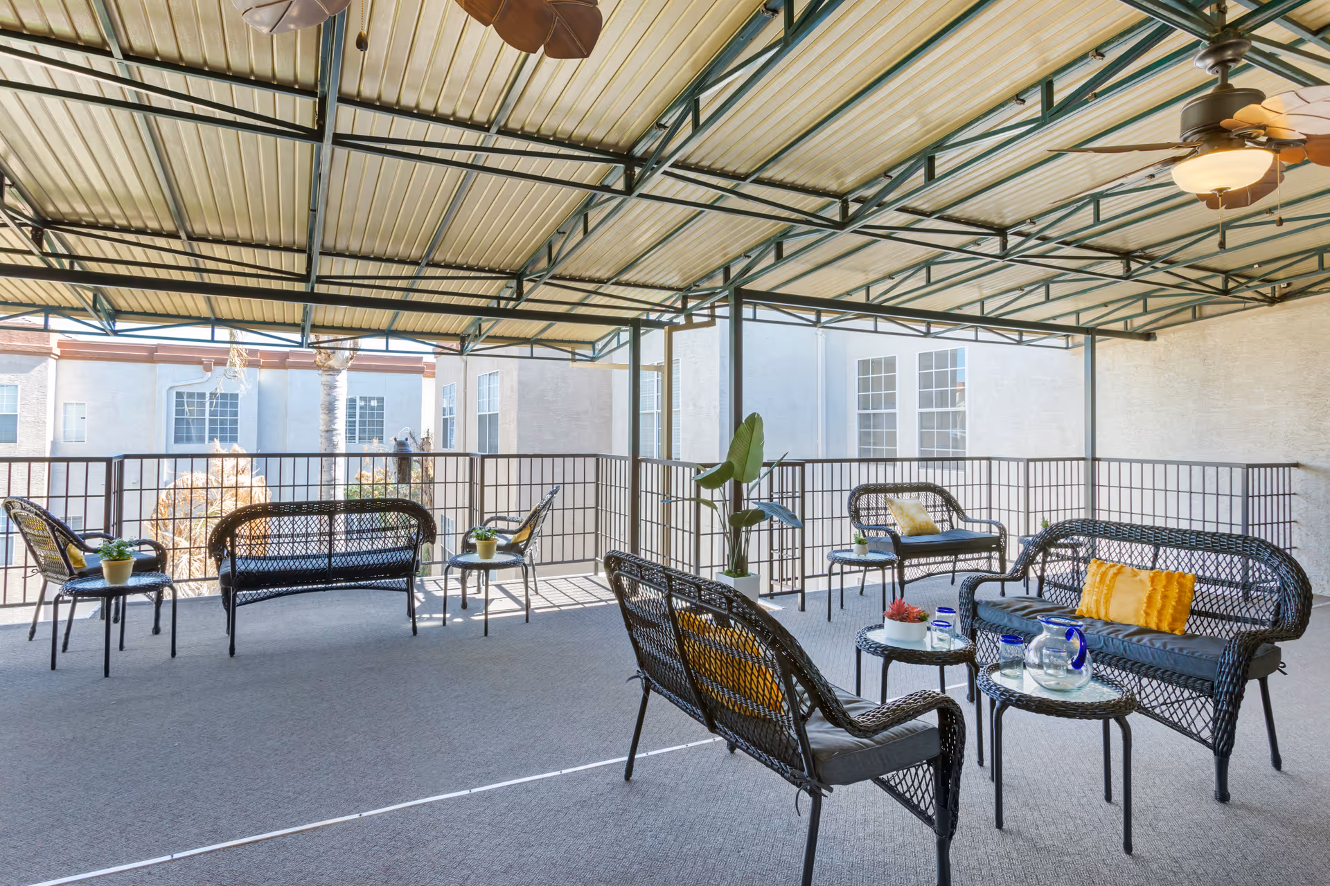 Covered outdoor patio area with black wicker seating including chairs and loveseats, small tables with potted plants and a glass pitcher with glasses, ceiling fans with lights, and surrounding buildings visible in the background.