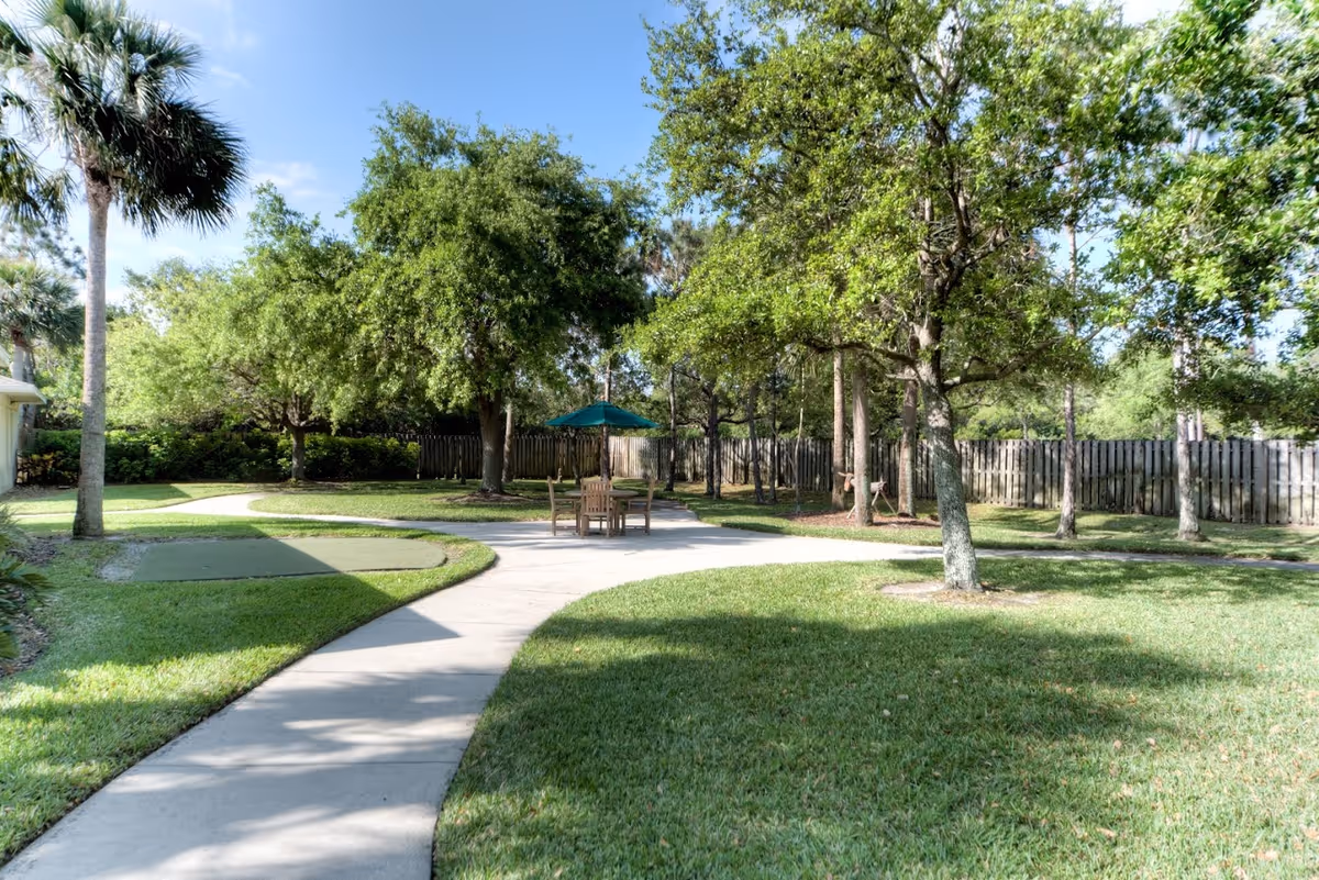 A sunny outdoor garden area with a curved concrete pathway, green grass, several trees, a wooden table with chairs, and a green umbrella. A wooden fence encloses the area.