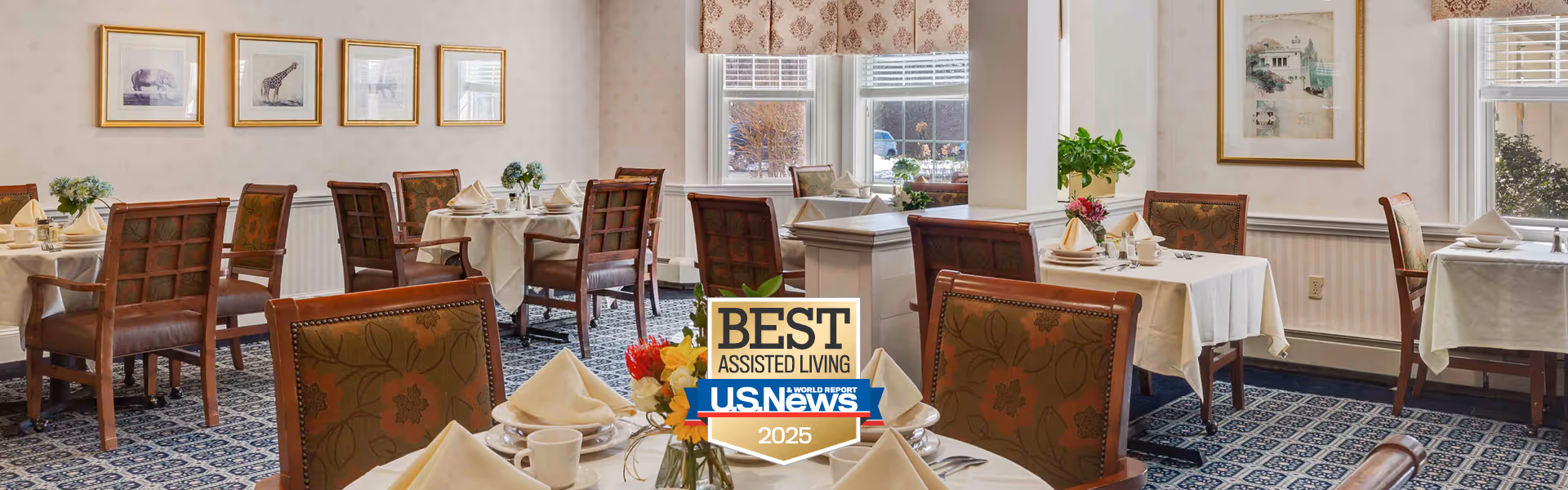 A bright and elegant dining room with several tables covered in white tablecloths, each set with napkins, cups, and small flower arrangements. The room features wooden chairs with patterned upholstery, framed artwork on the walls, and large windows letting in natural light.