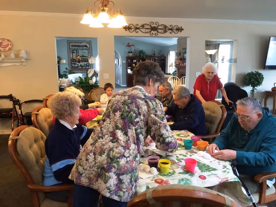 A group of elderly people sitting around a dining table engaged in an activity involving white eggs and colorful cups, with one woman standing and assisting them. The room is warmly lit with a chandelier overhead and decorated with plants and furniture.