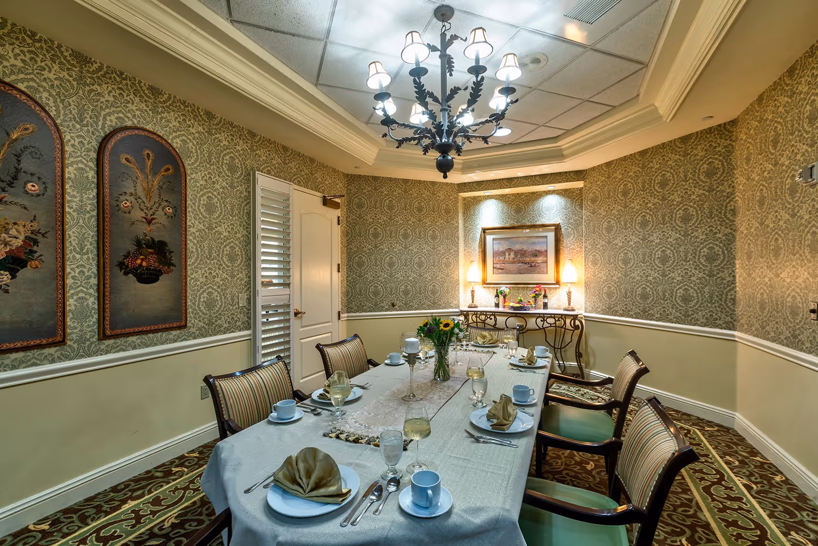 A formal dining room with a long table set for six people, featuring folded napkins, plates, cups, and glasses. The room has ornate wallpaper, a chandelier hanging from the ceiling, framed floral artwork on one wall, and a side table with lamps and a framed picture.