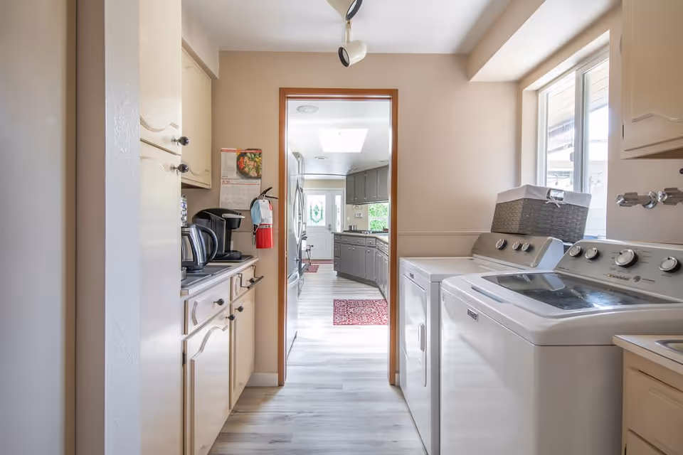 A bright laundry room with a washing machine and dryer on the right side, a window above them, and beige cabinets on the left side. The room leads into a kitchen area with gray cabinets and a refrigerator visible in the background. The floor is light-colored wood, and there is a red rug in the kitchen area.