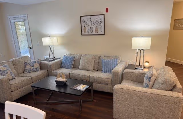 A cozy living room with a beige sofa, loveseat, and armchair arranged around a black coffee table. The sofas have blue and beige patterned cushions. Two table lamps with white shades are placed on side tables beside the sofa, and a framed artwork featuring vases with flowers hangs on the wall behind the sofa. The room has wooden flooring and a door with window blinds on the left side.