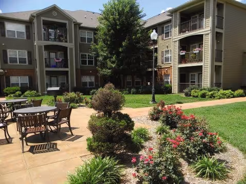 Outdoor courtyard area of a senior living facility with patio tables and chairs, landscaped garden beds with bushes and flowers, a paved walkway, and multi-story residential buildings in the background under a clear sky.