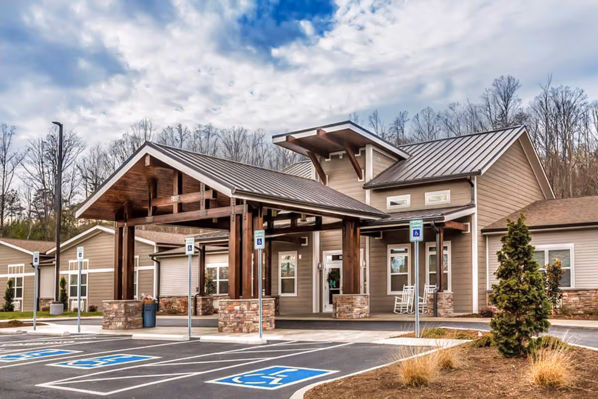 Exterior view of Pinnacle Assisted Living facility showing the main entrance with a covered drop-off area supported by wooden beams and stone pillars, surrounded by a parking lot with multiple handicap parking spaces and a few small trees and shrubs.