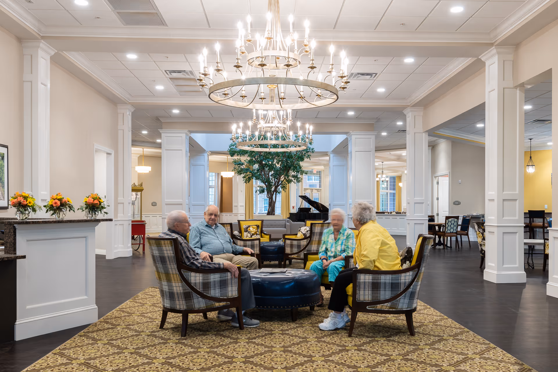 A spacious and well-lit common area in a senior living facility with four elderly people sitting and conversing on upholstered chairs arranged around a circular ottoman. The room features elegant chandeliers, white columns, a patterned carpet, and a grand piano in the background, with additional seating and tables visible.