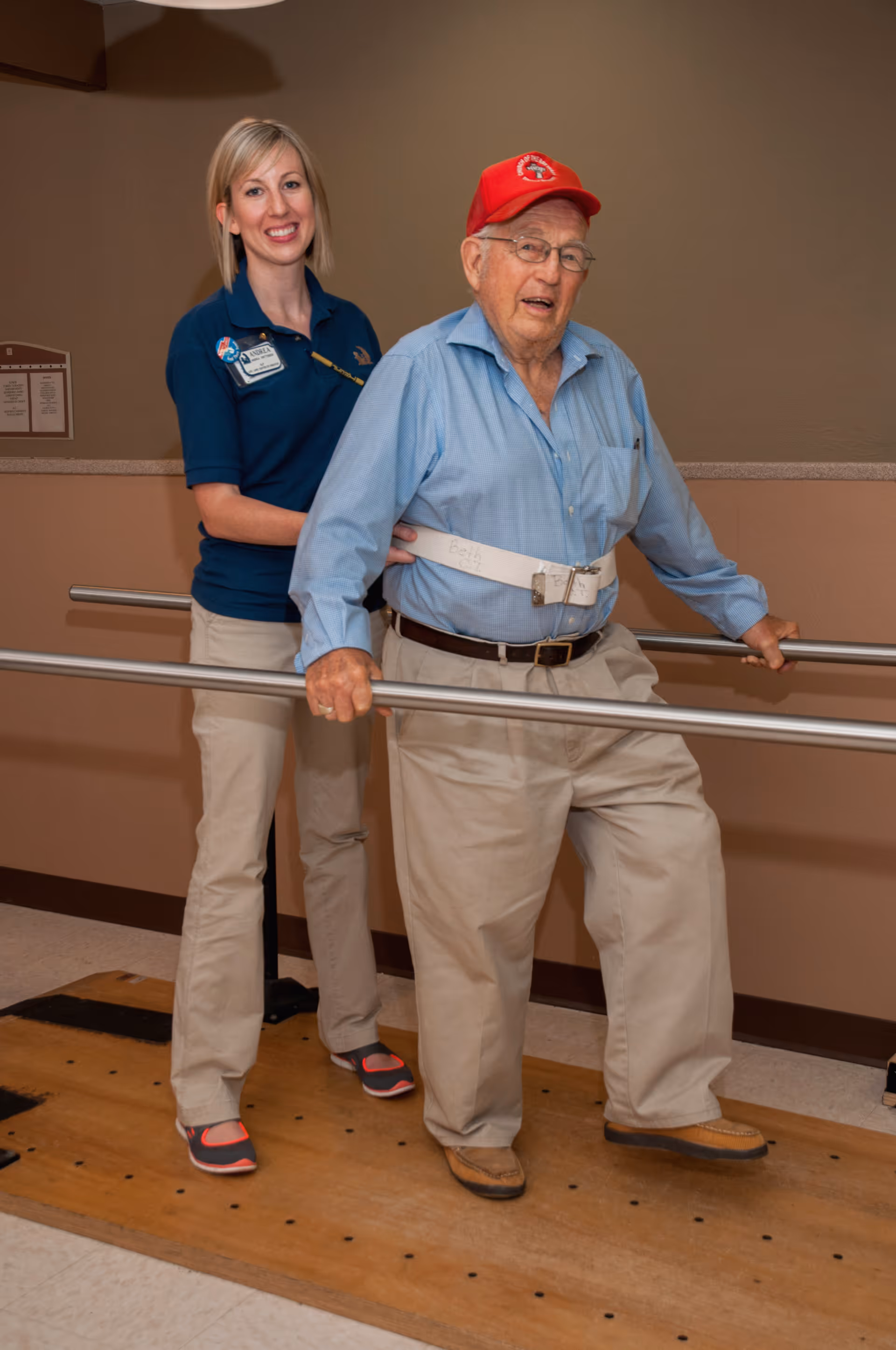 An elderly man wearing a red cap and light blue shirt is walking with support from parallel bars while a female caregiver in a blue shirt assists him from behind in a rehabilitation or therapy room.