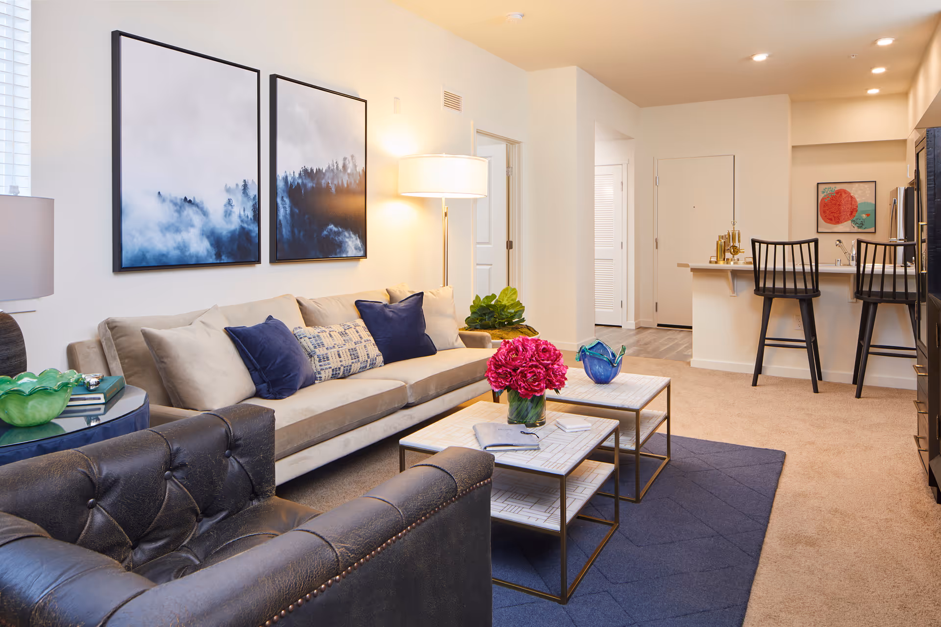 Modern furnished living room with a beige sofa, leather armchair, nested coffee tables topped with flowers, wall art, and a kitchen breakfast bar with stools.
