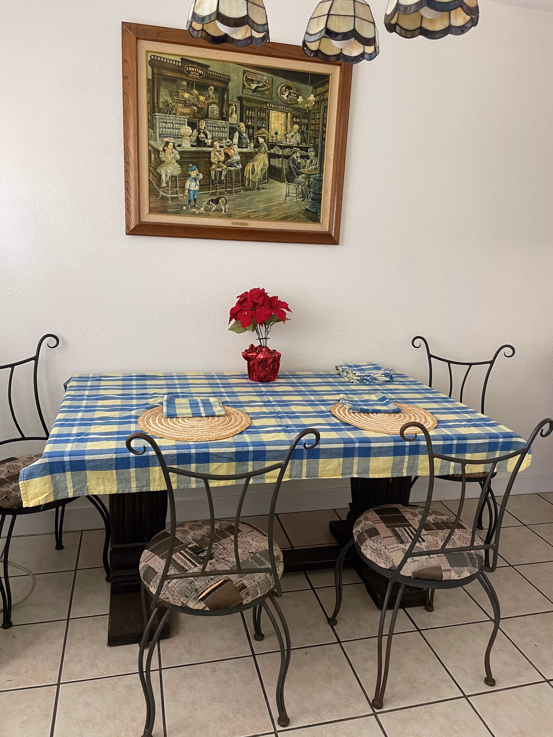A dining table covered with a blue and yellow checkered tablecloth, set with two round woven placemats and matching napkins. Four wrought iron chairs with patterned cushions surround the table. A small red poinsettia plant in a red foil wrapper is placed in the center of the table. A framed vintage-style painting depicting people in an old-fashioned store hangs on the white wall above the table. The floor is tiled with light-colored tiles.