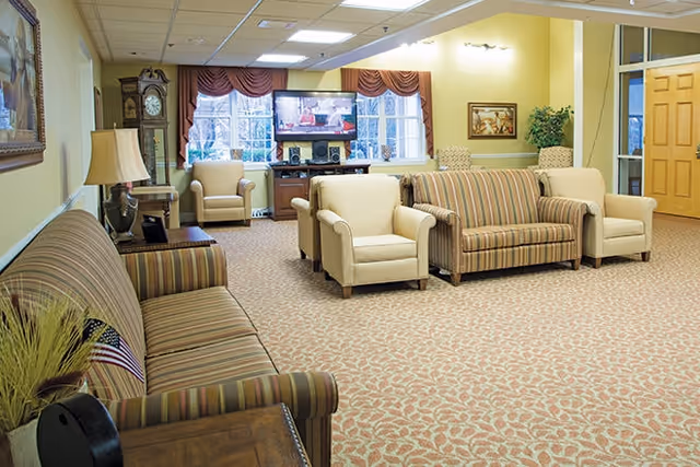 A spacious living room area with patterned carpet and multiple upholstered chairs and sofas arranged around the room. There is a large grandfather clock, a table lamp, and a flat-screen TV mounted on a stand near windows with red valances. The walls are painted light yellow and decorated with framed artwork and a potted plant. A wooden door is visible on the right side.