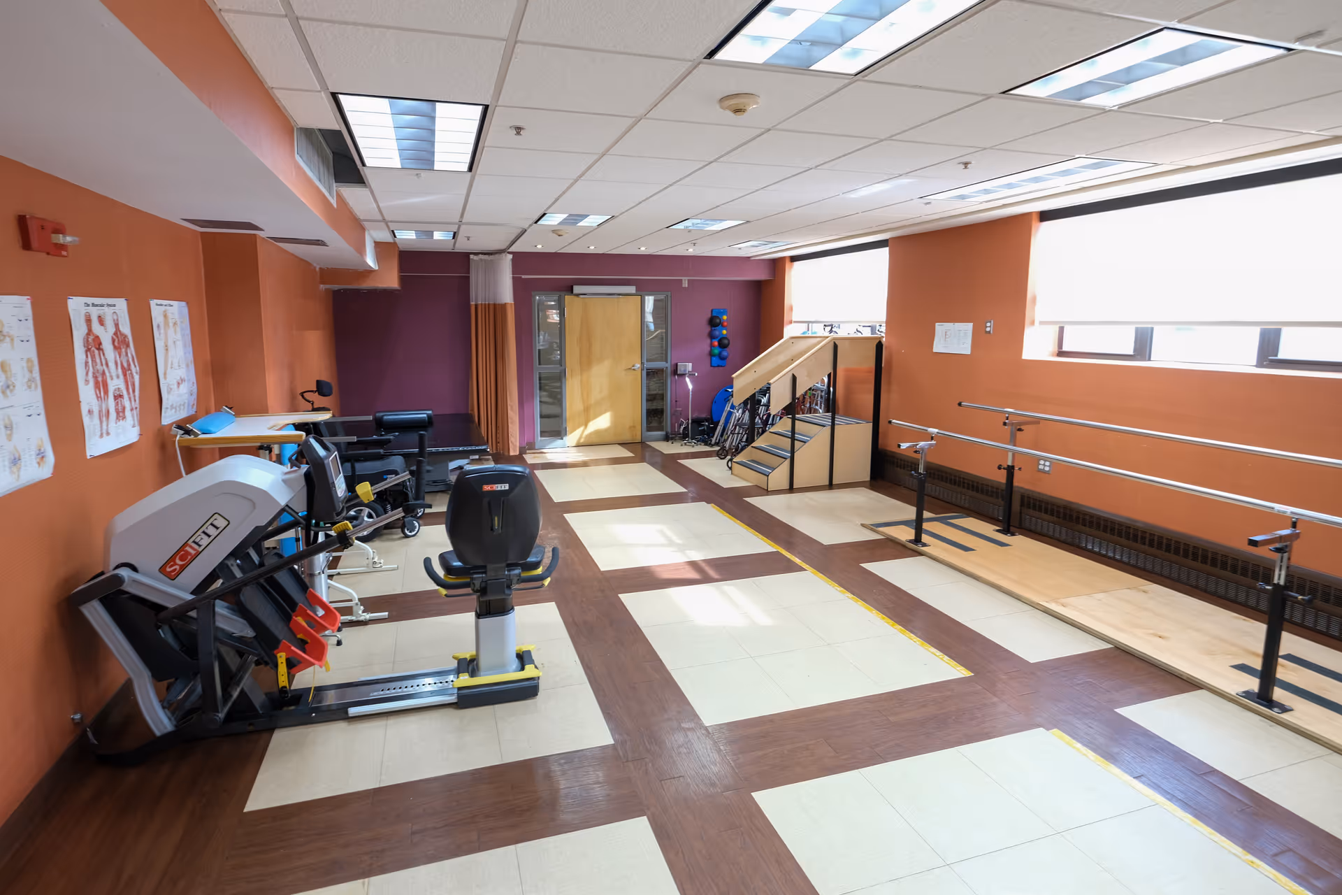 A rehabilitation or physical therapy room with exercise equipment including a recumbent bike, parallel bars for walking practice, a small staircase for step training, and anatomical charts on the wall. The room has orange and purple walls, large windows with blinds, and a tiled floor with a wood pattern.