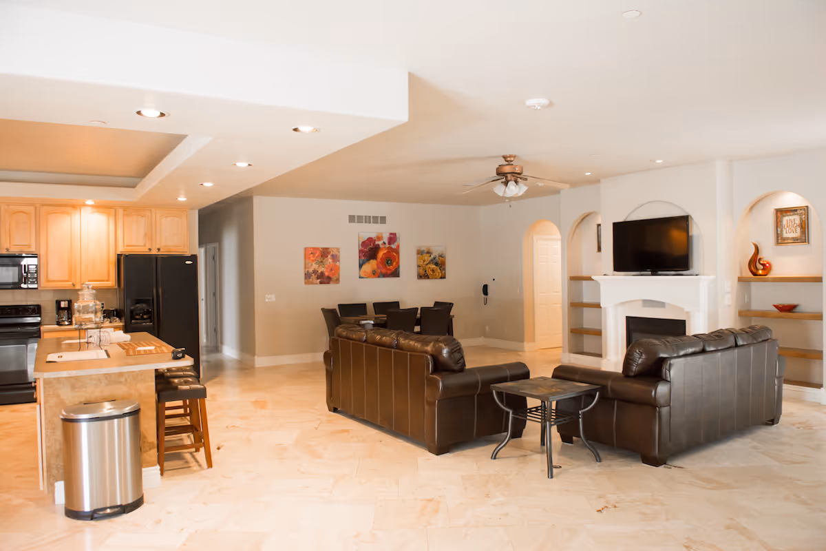 Open concept living area with a kitchen on the left featuring light wood cabinets, black appliances, and a kitchen island with stools. In the center, two brown leather sofas face a white fireplace with a mounted flat-screen TV above it. The room has light-colored tile flooring, recessed lighting, and decorative wall art and shelves.