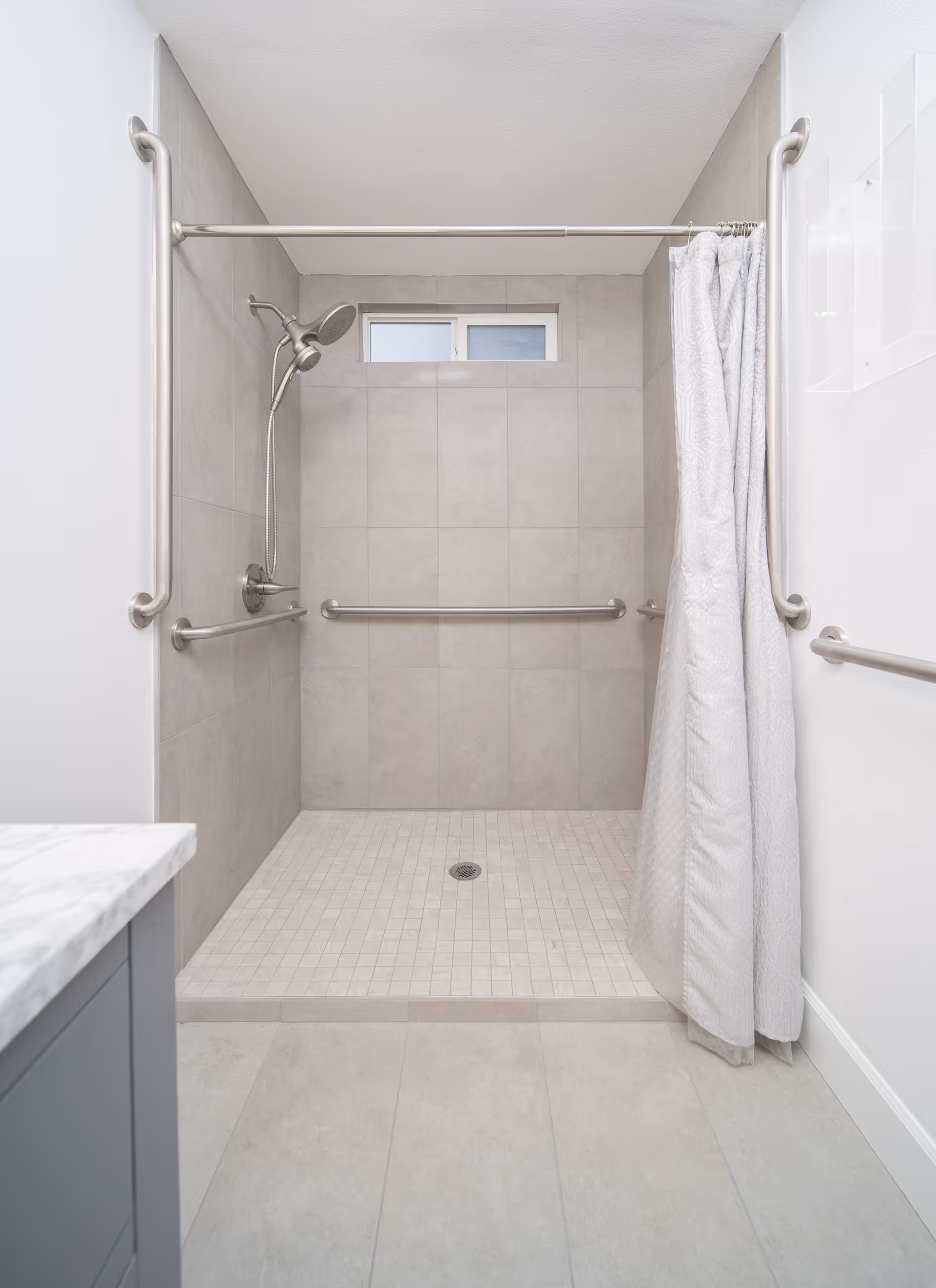 A walk-in shower with beige tiles on the walls and floor, equipped with multiple stainless steel grab bars for safety. There is a handheld showerhead mounted on the left wall and a white shower curtain partially drawn on the right side. A small frosted window is located above the shower area.