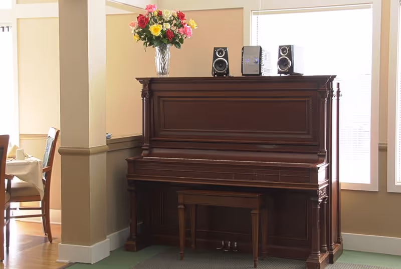 An upright wooden piano with a matching bench in front of two large windows. On top of the piano, there is a vase with colorful flowers and a small stereo system with two speakers. To the left, part of a dining area with chairs and a table covered with a white tablecloth is visible.