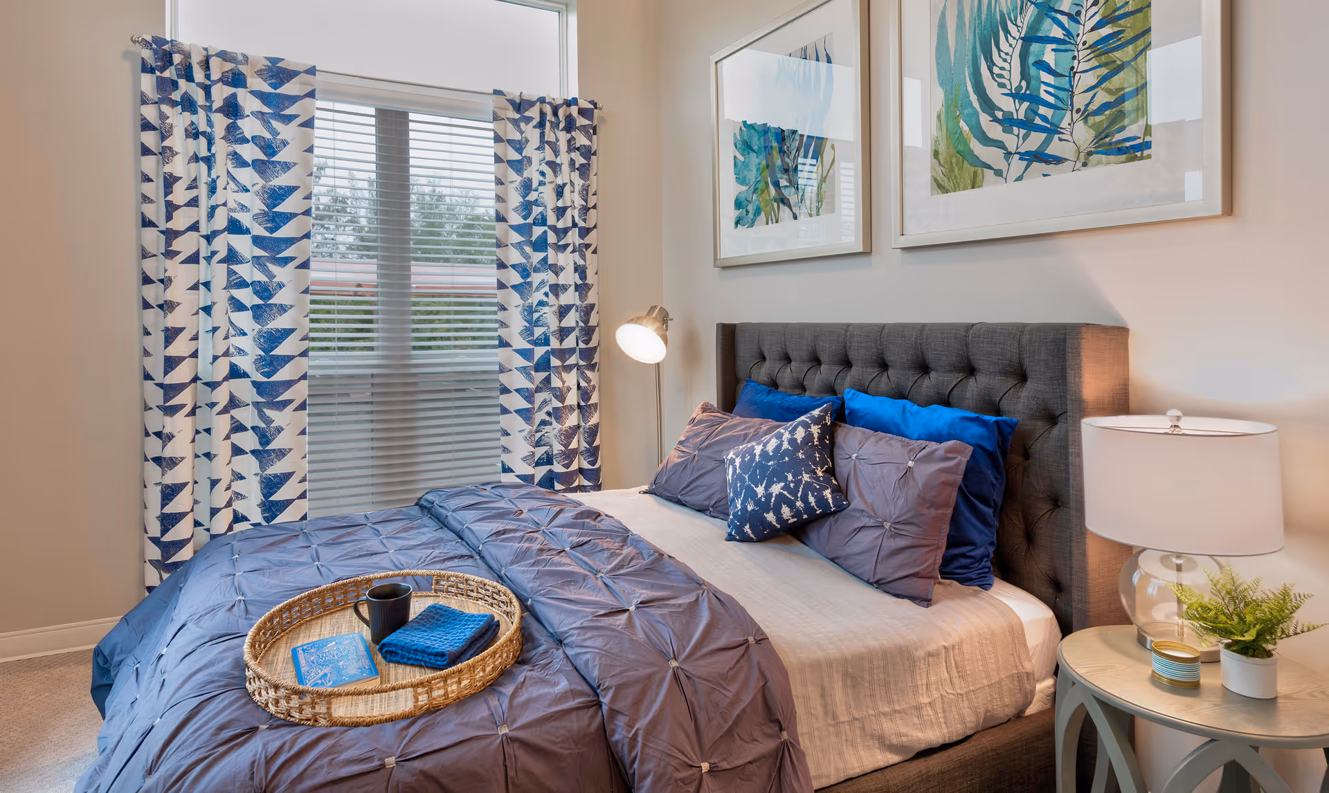 A neatly made bedroom with a tufted headboard, blue bedding and decorative pillows, patterned curtains over a window, and a nightstand with a lamp and plant.