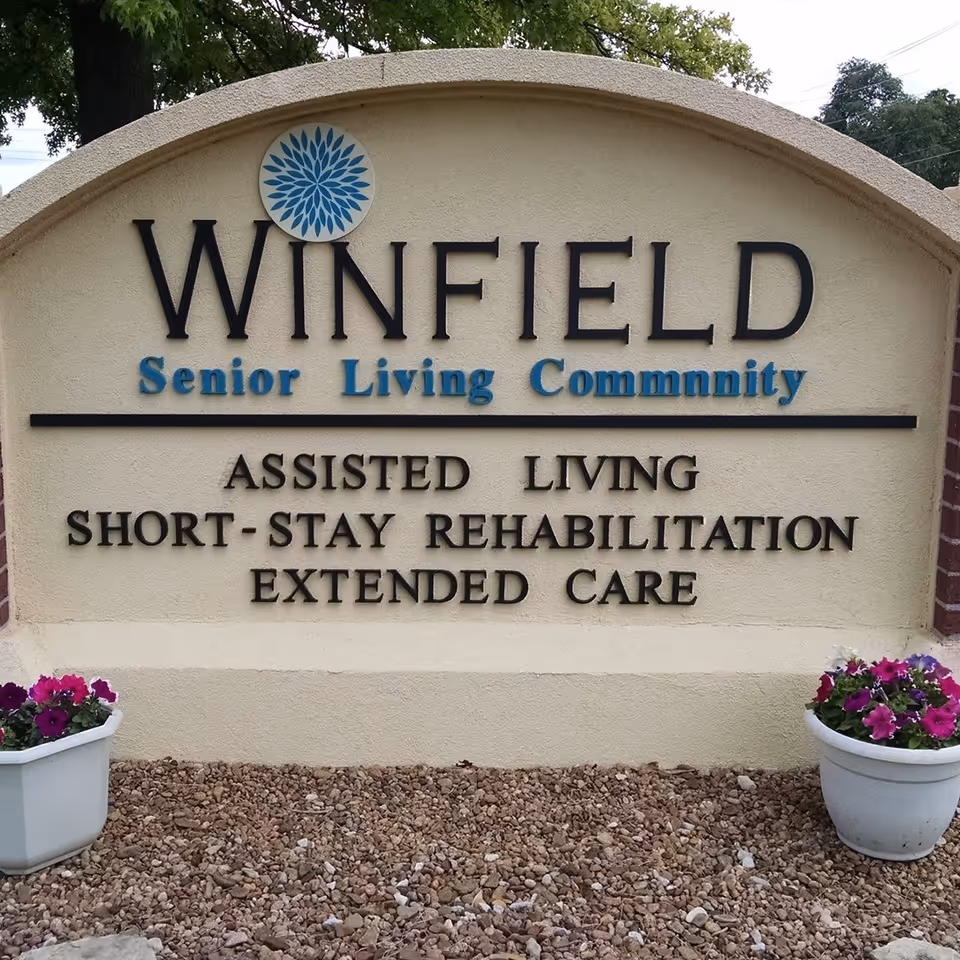 Outdoor sign for Winfield Senior Living Community with text indicating services including assisted living, short-stay rehabilitation, and extended care. The sign is beige with black and blue lettering, flanked by two white flower pots with purple and pink flowers, and set against a background of trees and sky.