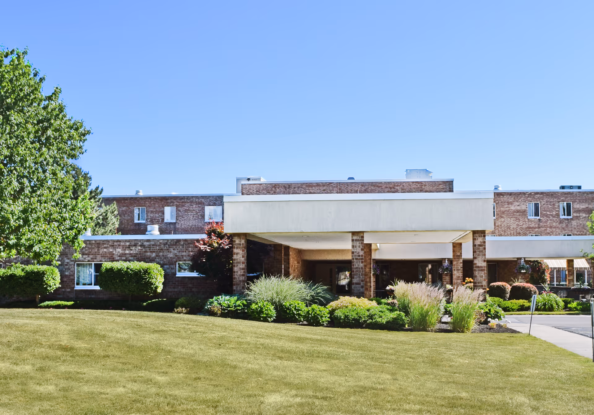 Exterior view of Elderwood at Hamburg senior living facility showing a brick building with a covered entrance, surrounded by green bushes, trees, and a well-maintained lawn under a clear blue sky.