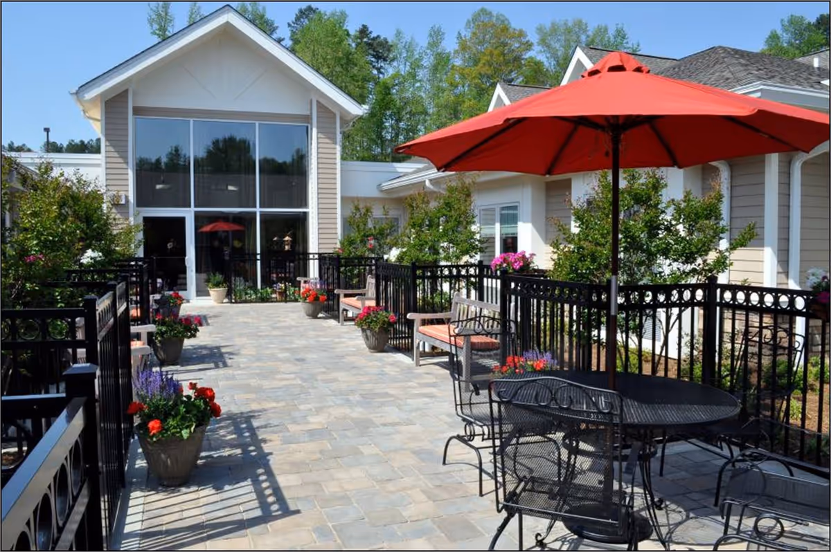 Outdoor patio area at Highland Hills featuring a paved walkway with black metal railings, several potted plants with colorful flowers, benches with cushions, and a black metal table with chairs under a large red umbrella. The patio is adjacent to a light-colored building with large windows and surrounded by greenery and trees.
