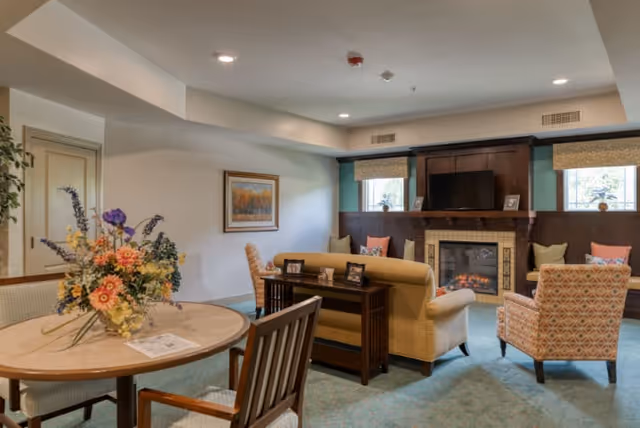 A cozy living room area with a fireplace and a mounted TV above it. The room features a beige sofa, patterned armchairs, and a wooden table with framed photos behind the sofa. There is a round table with a floral arrangement and chairs in the foreground. Two windows with blinds flank the fireplace, and a framed painting hangs on the wall.