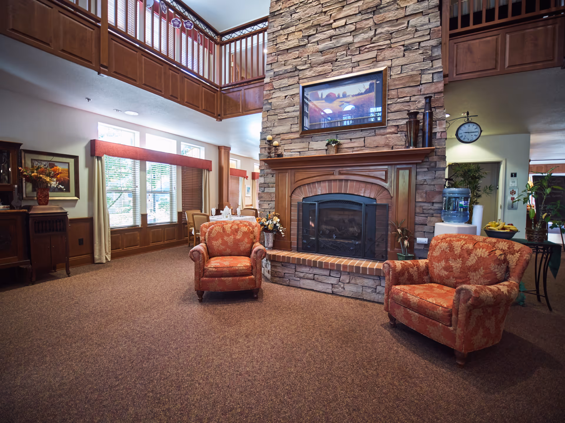 Cozy common room with two patterned armchairs facing a large stone fireplace beneath a second-floor balcony.