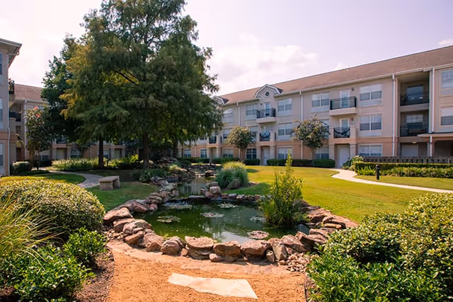 A landscaped outdoor area at Brookdale Clear Lake featuring a small pond surrounded by rocks, green bushes, and trees. The background shows a multi-story residential building with balconies and windows under a partly cloudy sky.