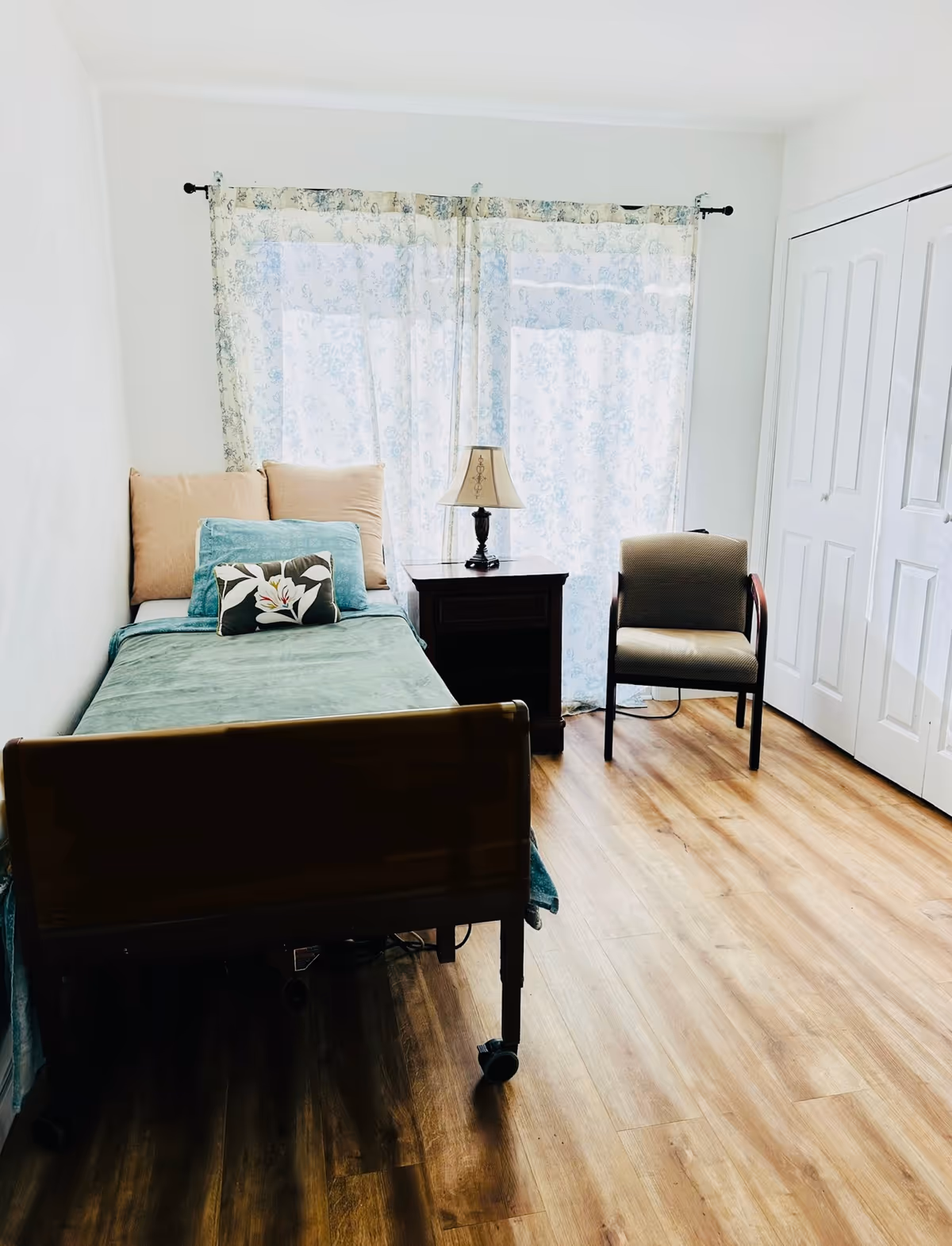A simple bedroom with a single bed covered in green bedding and multiple pillows, a wooden nightstand with a lamp, a cushioned chair, and a closet with white double doors. The room has light-colored walls, wood flooring, and sheer floral curtains covering a window.