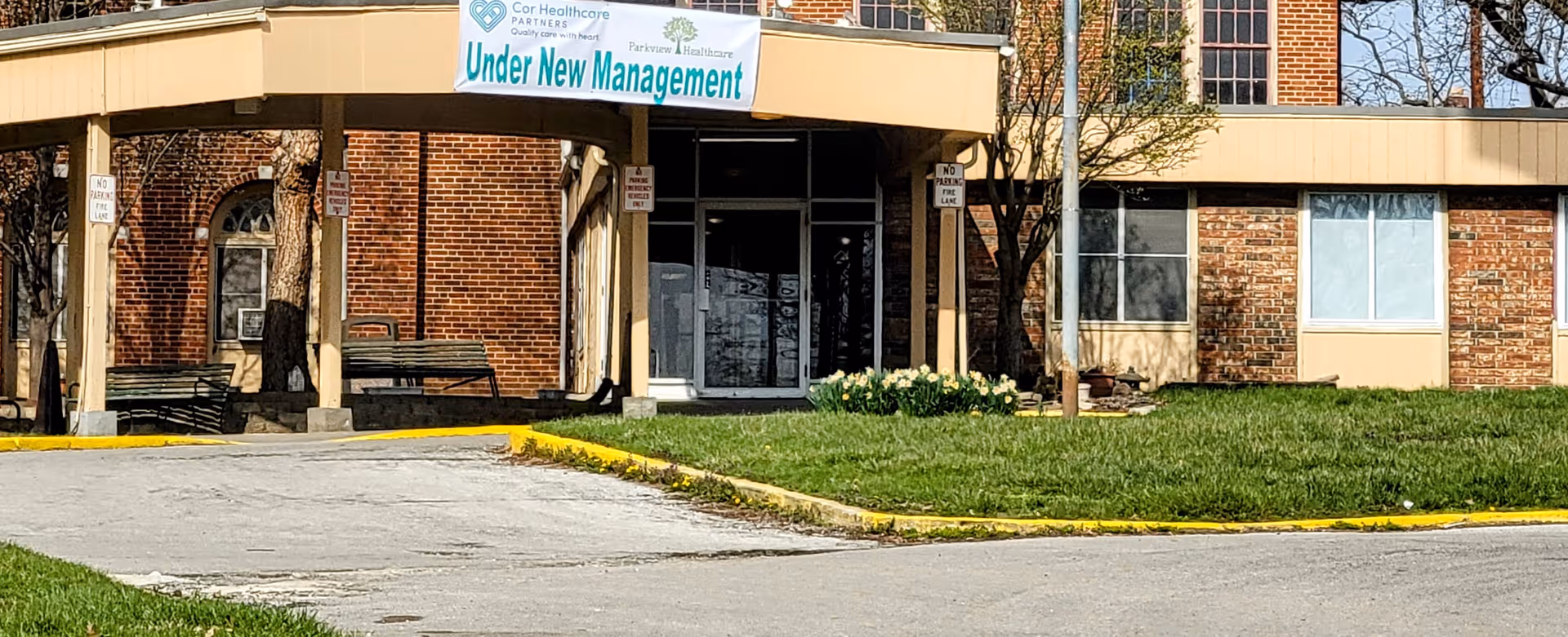 Front entrance of a brick senior living facility with a covered drive and a banner reading "Under New Management".