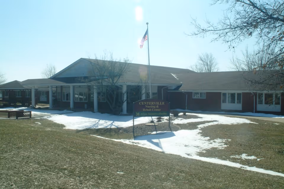 Exterior view of a single-story brick building with a covered entrance supported by white columns. There is a flagpole with an American flag in front of the building, and a sign that reads 'Centerville Nursing & Rehab Center.' The ground has patches of snow and grass, and there are leafless trees around the building.