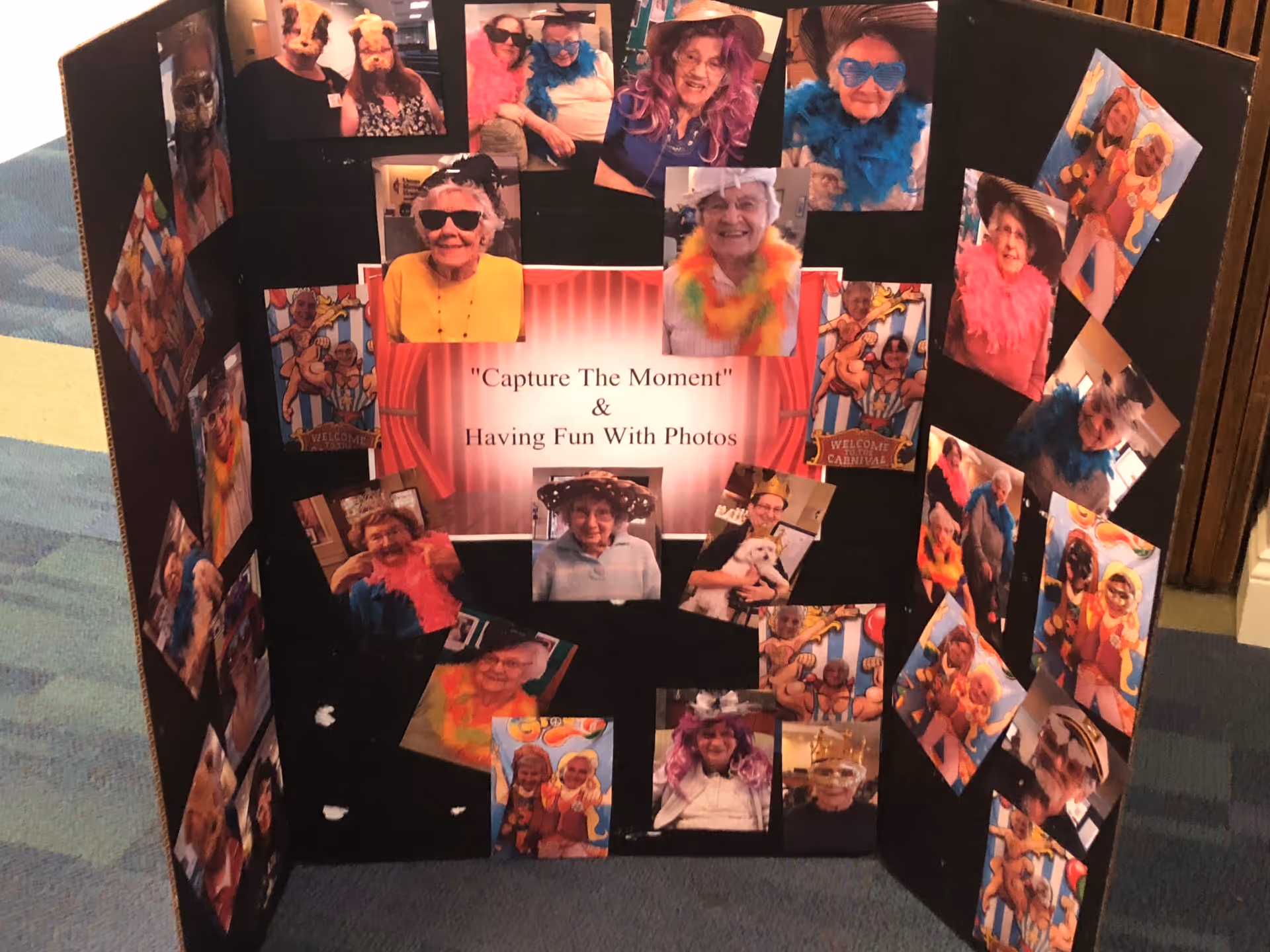 A black tri-fold display board featuring multiple photos of elderly individuals wearing colorful costumes, hats, sunglasses, and feather boas. The center of the board has a sign that reads 'Capture The Moment & Having Fun With Photos.' The display is set on a carpeted floor with a striped pattern.