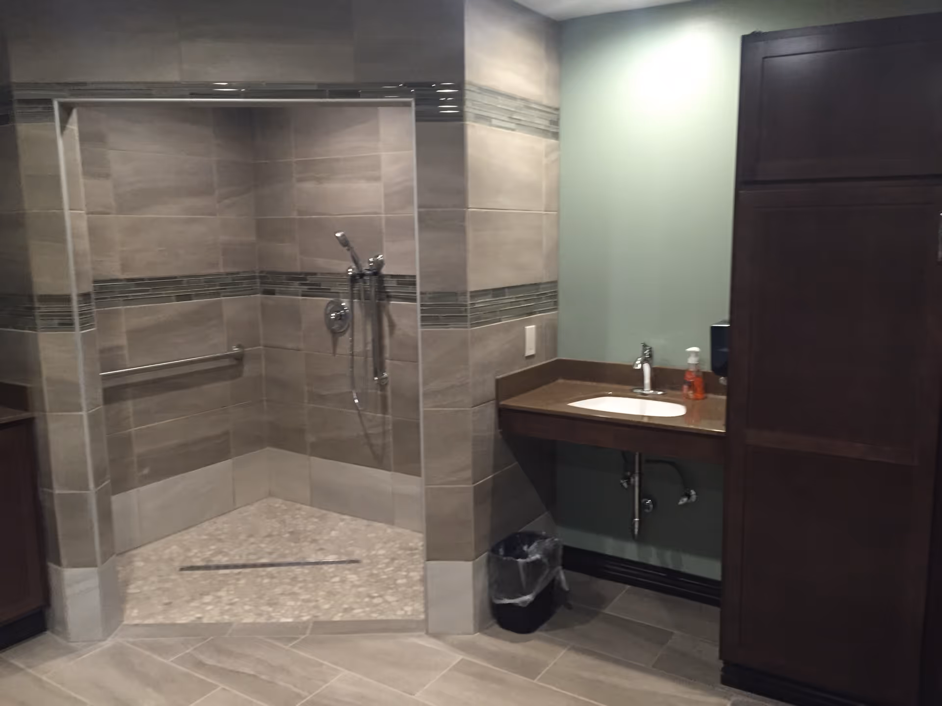 A modern bathroom featuring a walk-in shower with beige and gray tiles, a handheld showerhead, and a grab bar. To the right is a countertop with a sink, a soap dispenser, and a trash bin underneath. There is a tall wooden cabinet next to the sink area.