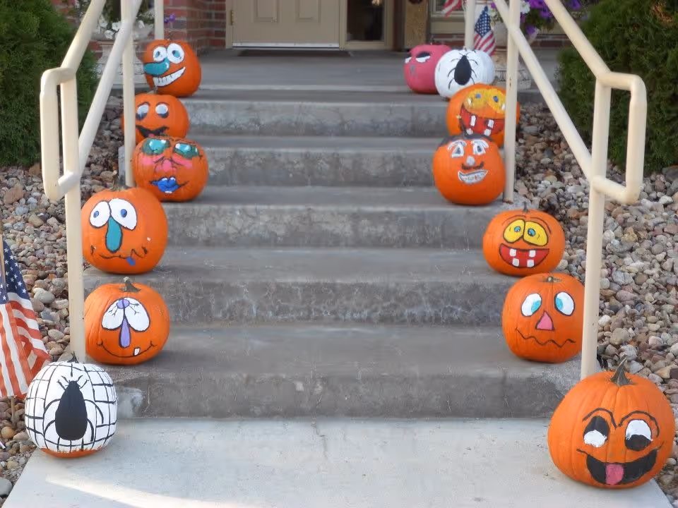 Concrete steps leading to a building entrance decorated with various painted pumpkins featuring different facial expressions and designs on both sides of the stairs. There are small American flags placed near the pumpkins and rocks lining the sides of the steps.