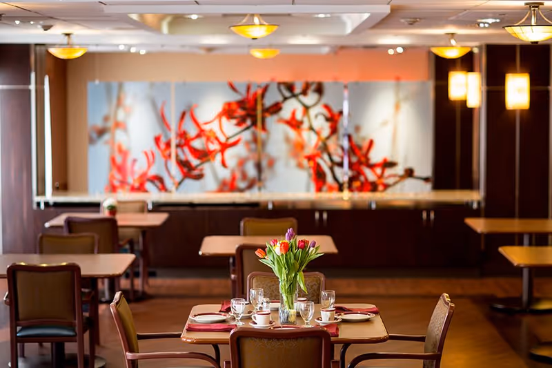 A dining room with several tables and chairs. One table in the foreground is set with plates, cups, glasses, and a vase of colorful tulips. The background features a large decorative panel with red and orange floral artwork and warm overhead lighting.