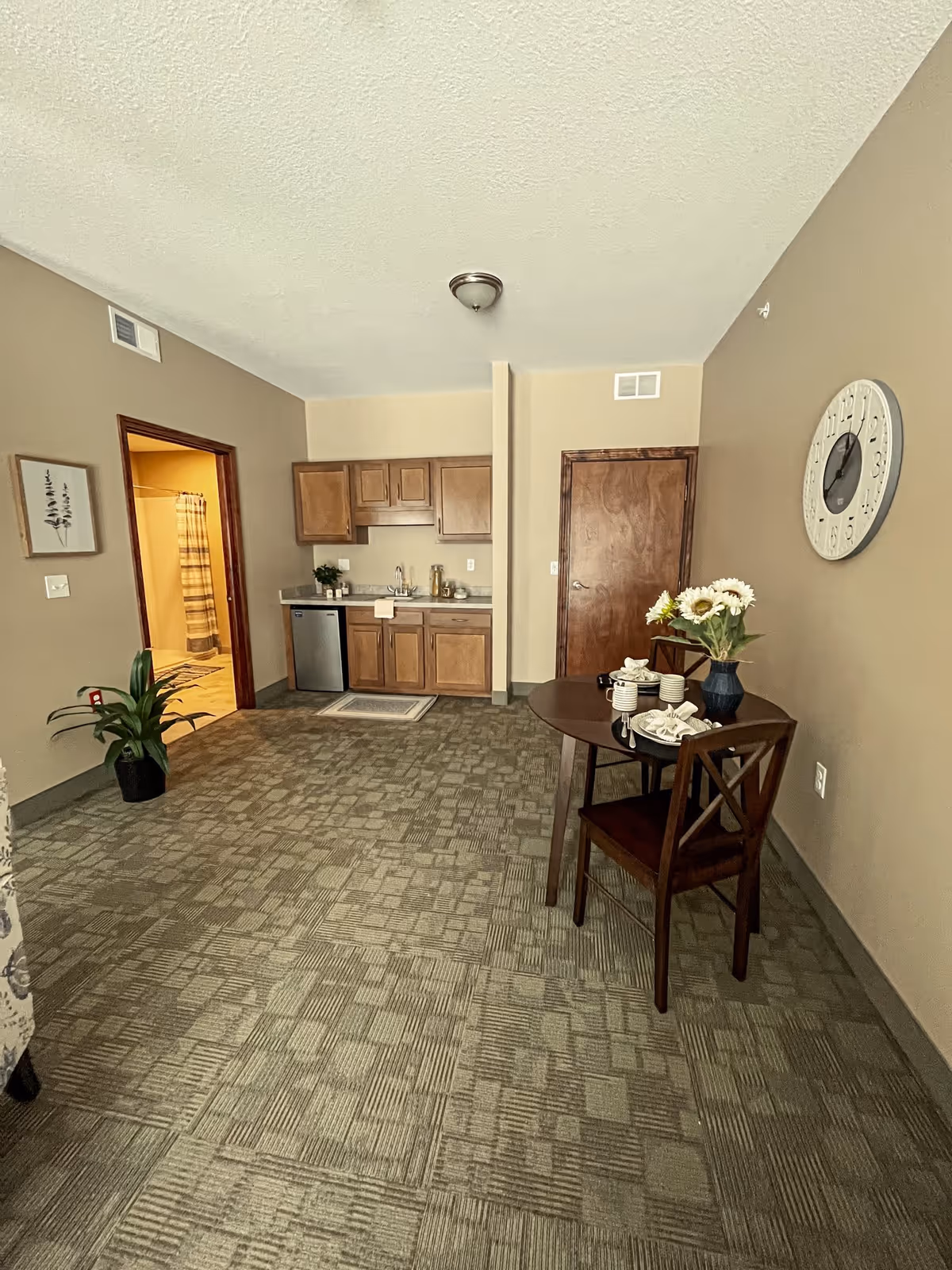 Interior view of a room with beige walls and patterned carpet. There is a small wooden dining table with two chairs, set with cups, plates, and a vase of flowers. On the right wall, there is a large round clock. The back wall features a kitchenette with wooden cabinets, a small refrigerator, and a sink. To the left, there is an open doorway leading to a bathroom with a shower curtain visible. A potted plant is placed near the doorway.