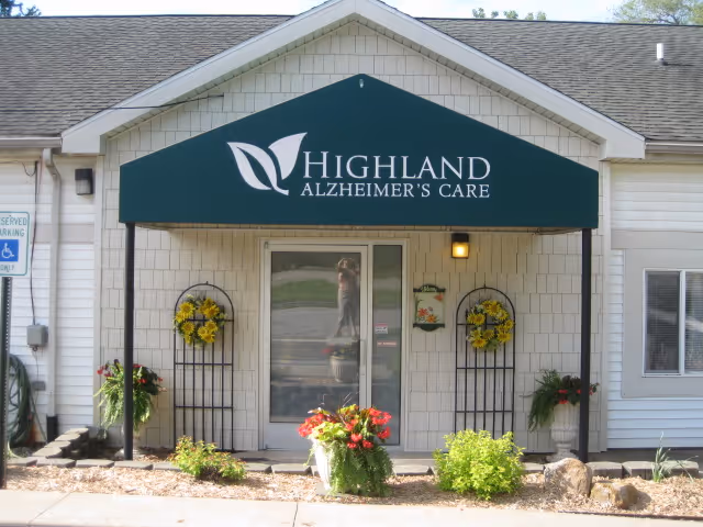 Entrance to Highland Alzheimer's Care facility with a green awning displaying the facility name. The entrance has glass double doors flanked by decorative flower arrangements and plants in front of a white shingled building.