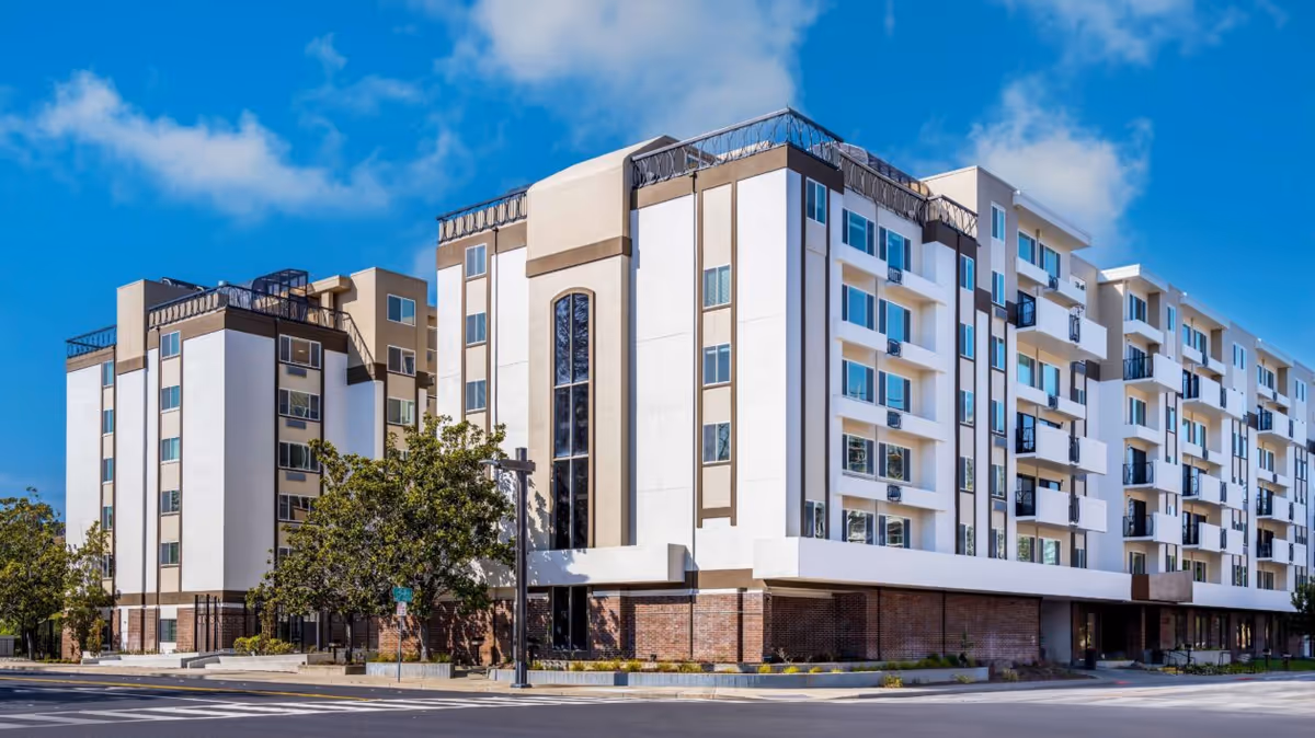 Exterior view of a multi-story residential building with white and beige walls, multiple windows, and balconies under a blue sky with some clouds. Trees and a street with a crosswalk are visible in front of the building.