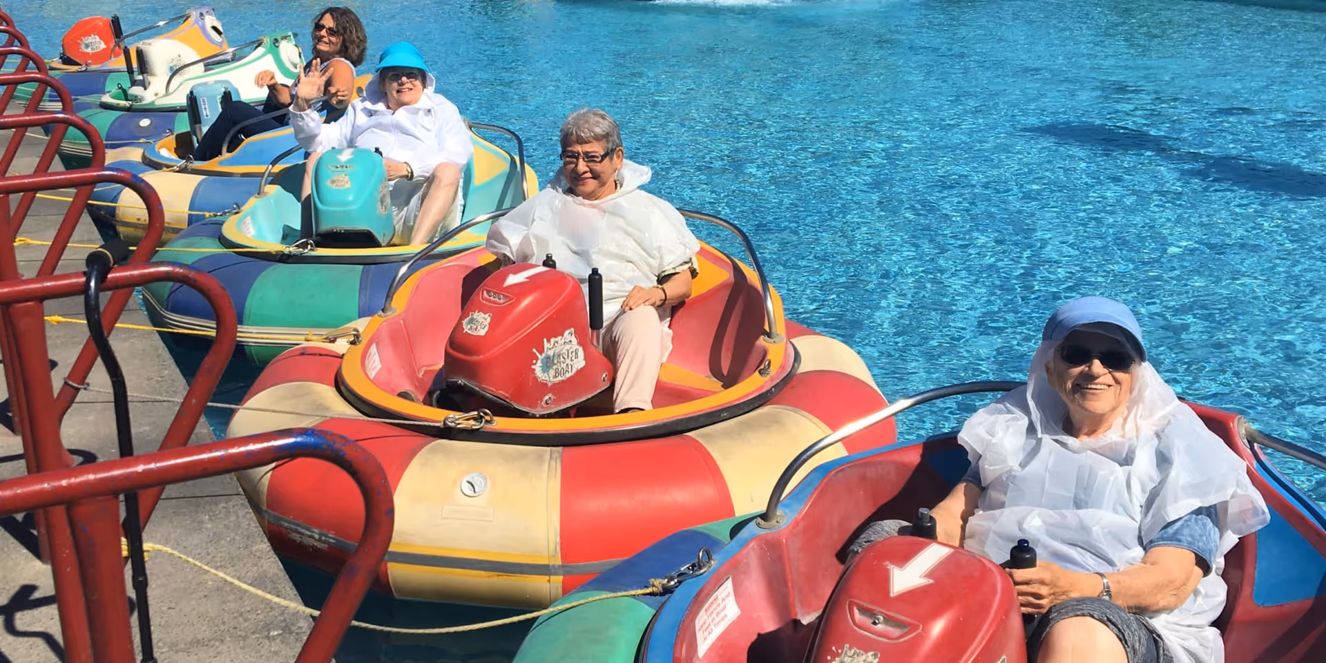 Four elderly women sitting in colorful bumper boats on a pool, smiling and waving, enjoying a sunny day outdoors.