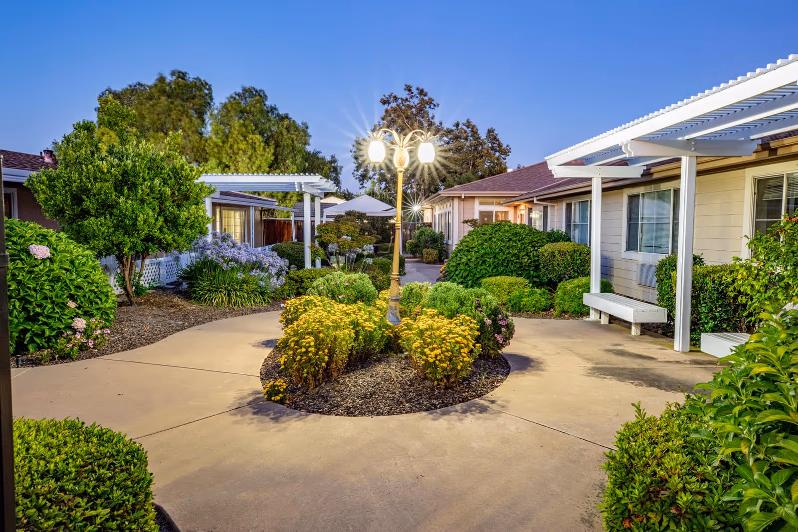 Outdoor courtyard area at Tuscany Villa Senior Living featuring a central lamp post with three glowing lights surrounded by yellow flowers and various green shrubs. The courtyard is paved with a walkway that splits around the flower bed, with single-story buildings and white pergolas on either side. Trees and additional plants are visible in the background under a clear evening sky.