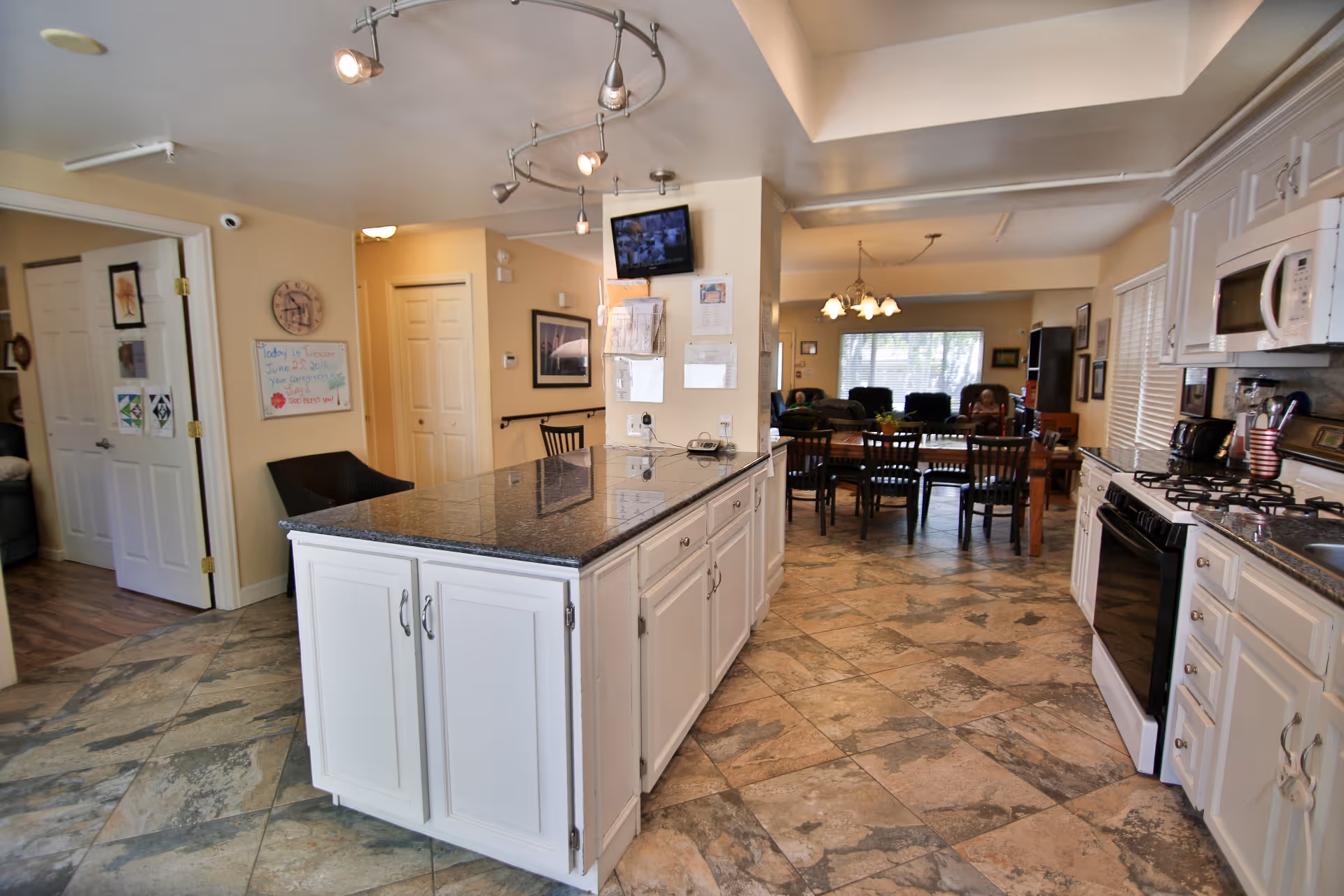 Interior view of a senior living facility showing a kitchen area with white cabinets, a granite countertop island, a stove, microwave, and coffee maker. Beyond the kitchen is a dining area with a wooden table and chairs, and a living room with armchairs and a large window letting in natural light.