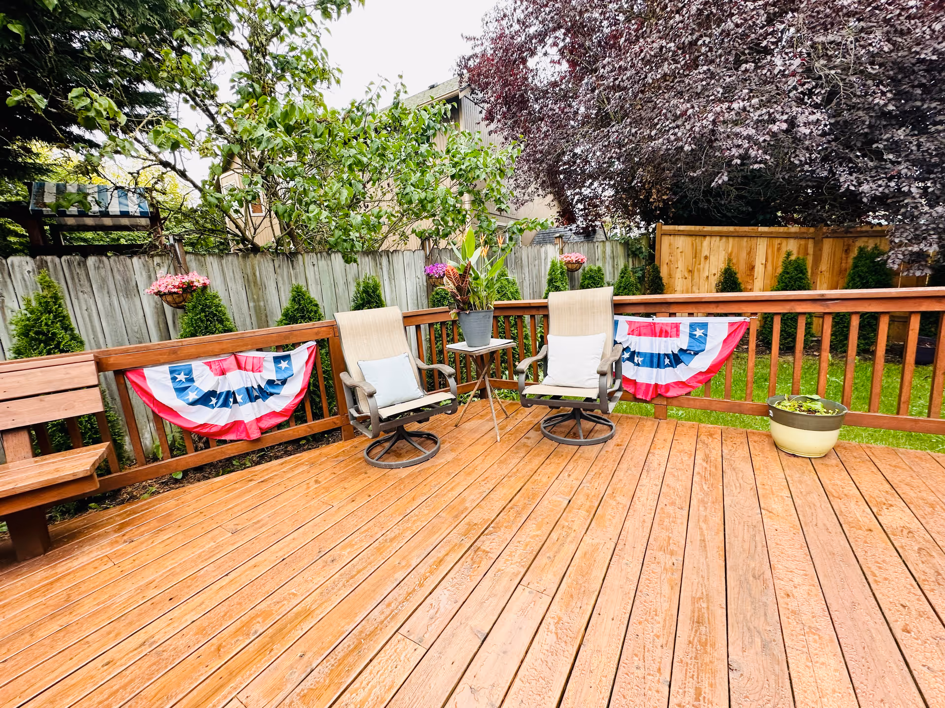 A wooden outdoor deck with two cushioned swivel chairs and a small table with a potted plant between them. The deck railing is decorated with red, white, and blue patriotic bunting. There are hanging flower pots and a wooden fence surrounding the yard with green grass and trees in the background.