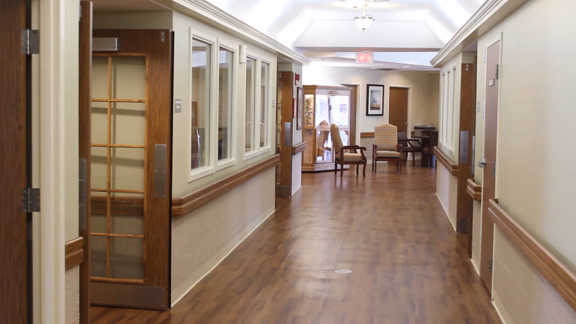 A well-lit hallway in a senior living facility with wooden flooring and beige walls. There are wooden handrails along both sides of the hallway, several closed doors, and a few chairs arranged near the end of the corridor under a skylight ceiling.