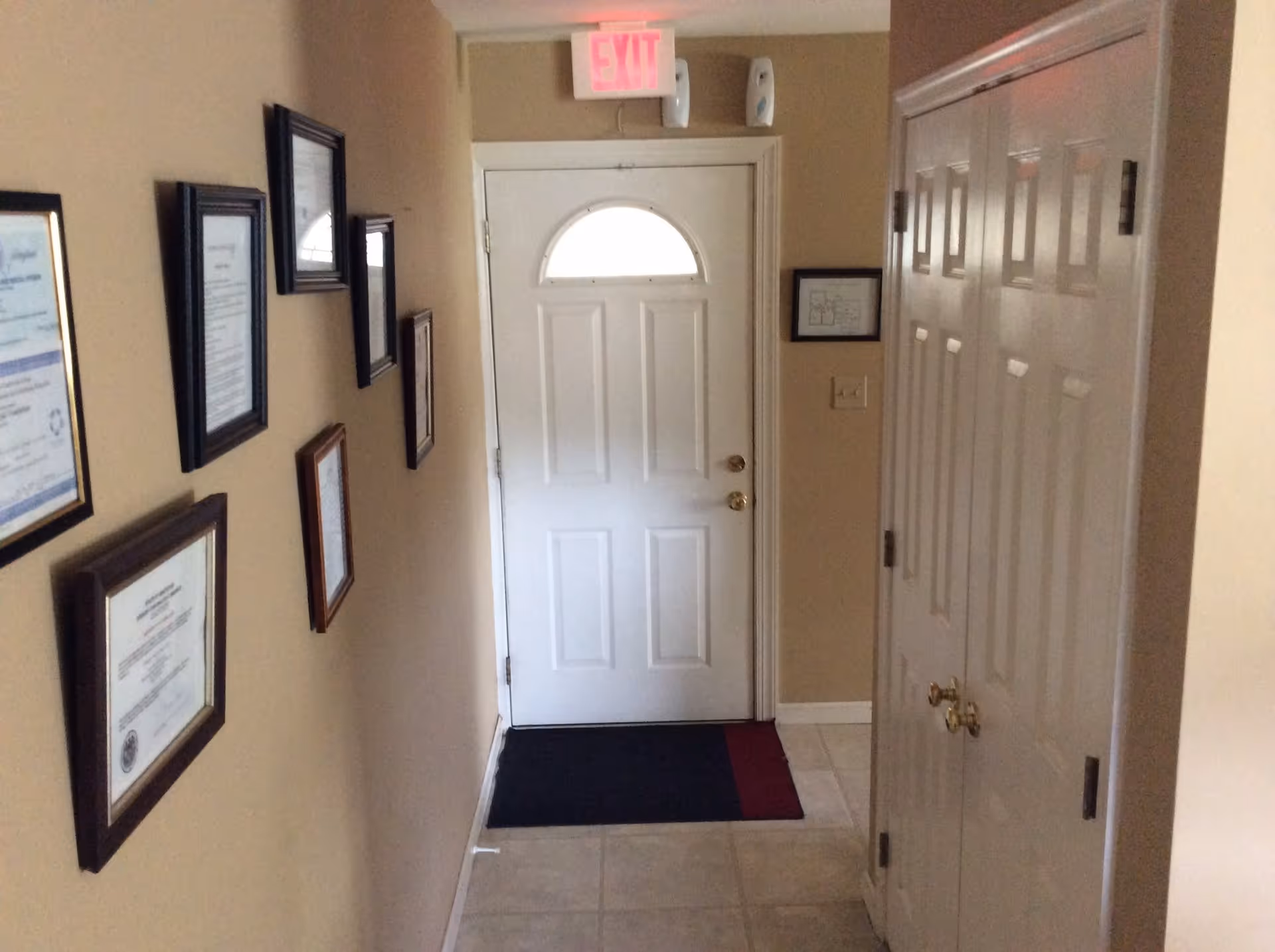 Narrow interior entry hallway with a white front door, framed certificates on the left wall, and closet doors on the right.