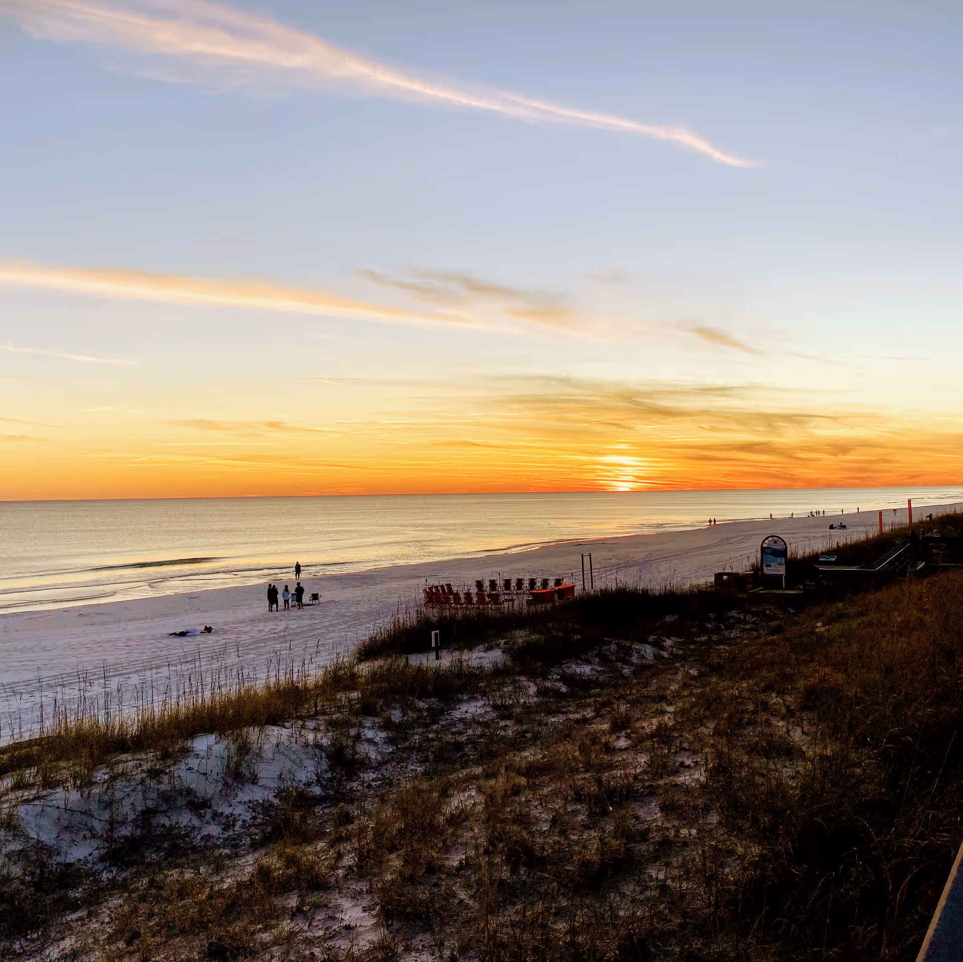 Sunset view over a sandy beach with a few people walking and standing near the shoreline. There are beach chairs arranged in a circle and some grassy dunes in the foreground.