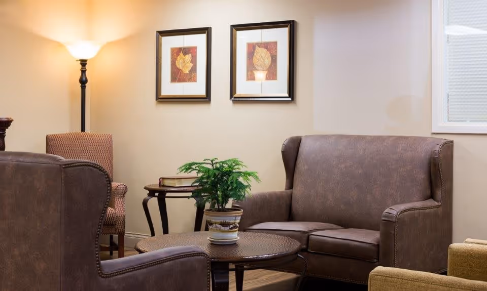 A cozy living room area with brown leather armchairs and a matching loveseat. There is a round coffee table with a potted plant on it, a side table with books, a floor lamp, and two framed leaf artworks on the beige wall.