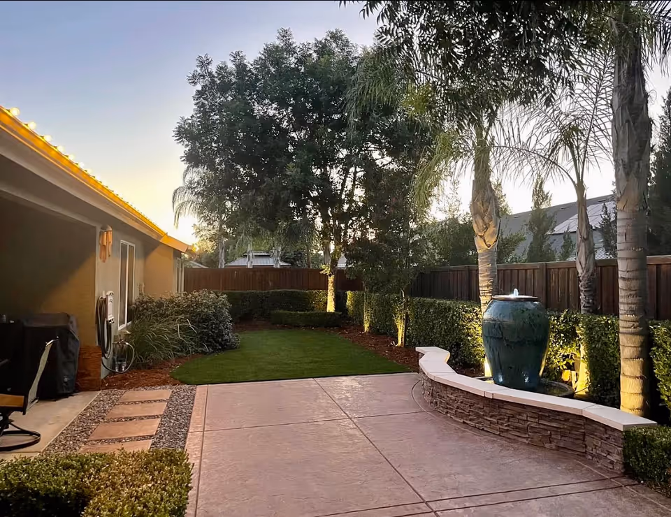 A backyard patio area at dusk with a concrete floor, a curved stone bench, a large blue water fountain, palm trees, and other greenery including bushes and a tree. The side of a house with outdoor lighting and a covered grill area is visible on the left.