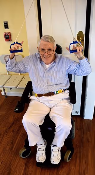 An elderly man sitting in a wheelchair indoors, smiling and holding up blue exercise handles attached to cords, wearing a striped shirt, white pants, and white sneakers.