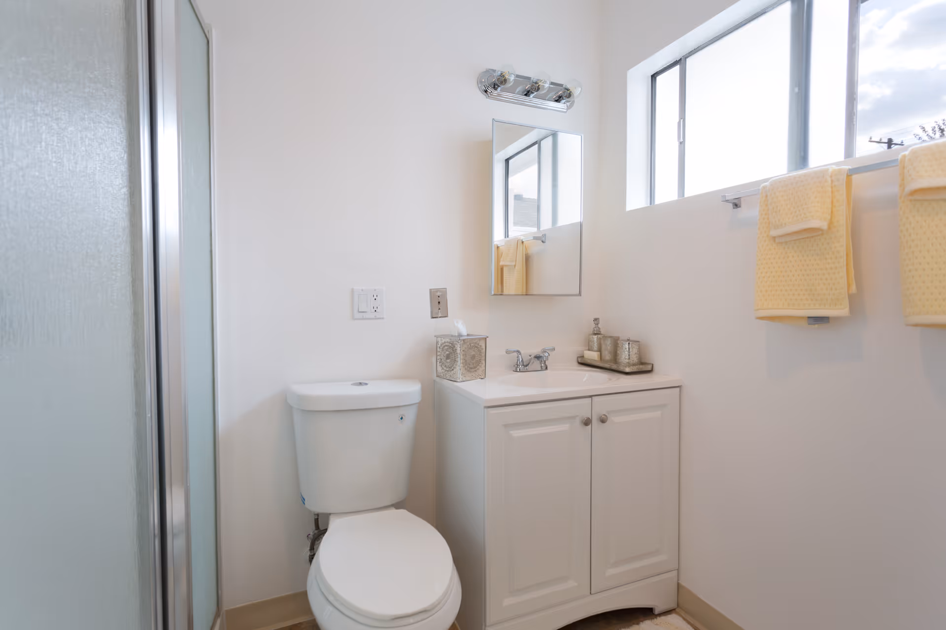 A clean bathroom with a white toilet, a white vanity with a sink, a rectangular mirror above the sink, and a window with two yellow towels hanging on a towel rack. There is a tissue box and decorative items on the vanity, and a shower with a frosted glass door is partially visible on the left.
