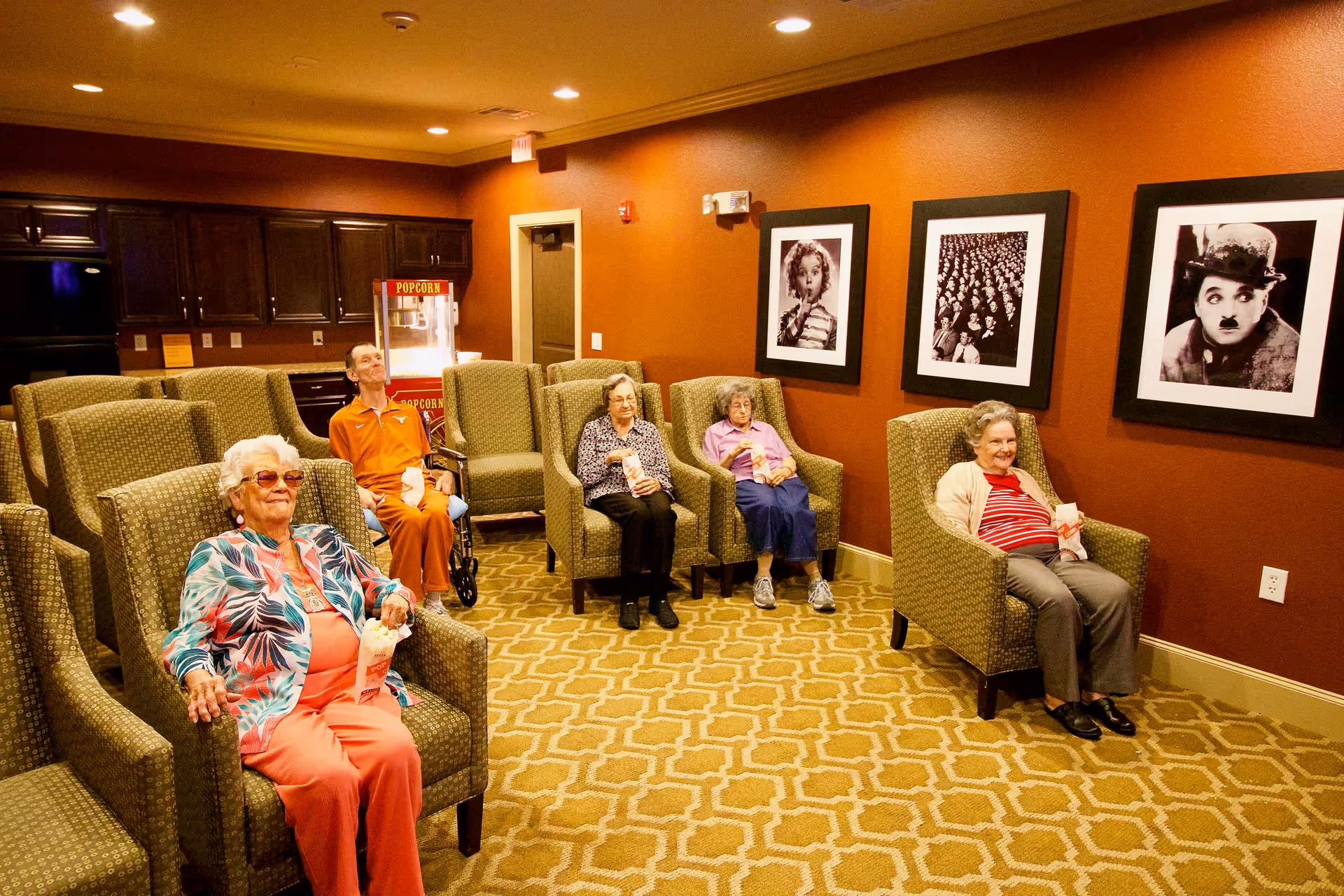 A group of elderly people sitting in comfortable armchairs in a cozy room with warm lighting and orange walls. They are holding popcorn bags, suggesting they are watching a movie or enjoying a social event. The room has framed black and white pictures on the wall and a popcorn machine in the background.